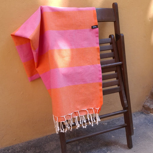 Fouta telo mare Foutalìa a righe rosa e arancione appesa a una sedia pieghevole di legno chiusa, all'aperto, con le frange bianche e luce solare naturale su parete ocra.