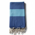 Telo mare Fouta in cotone riciclato blu e azzurro a righe grandi, piegato verticalmente su sfondo bianco con frange bianche.