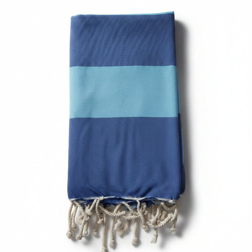 Telo mare Fouta in cotone riciclato blu e azzurro a righe grandi, piegato verticalmente su sfondo bianco con frange bianche.