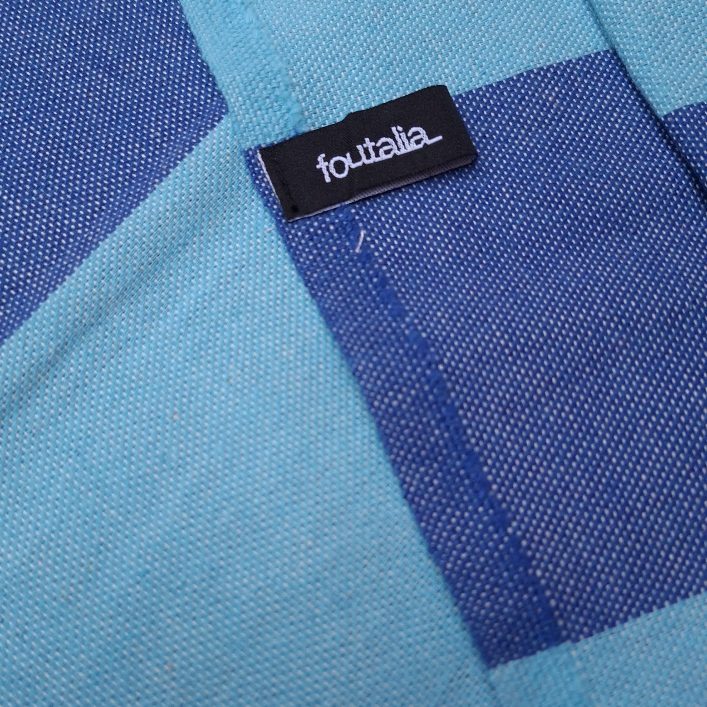 Primo piano dell'etichetta nera in tessuto con il brand foutalia, cucita sul bordo del telo mare blu.
