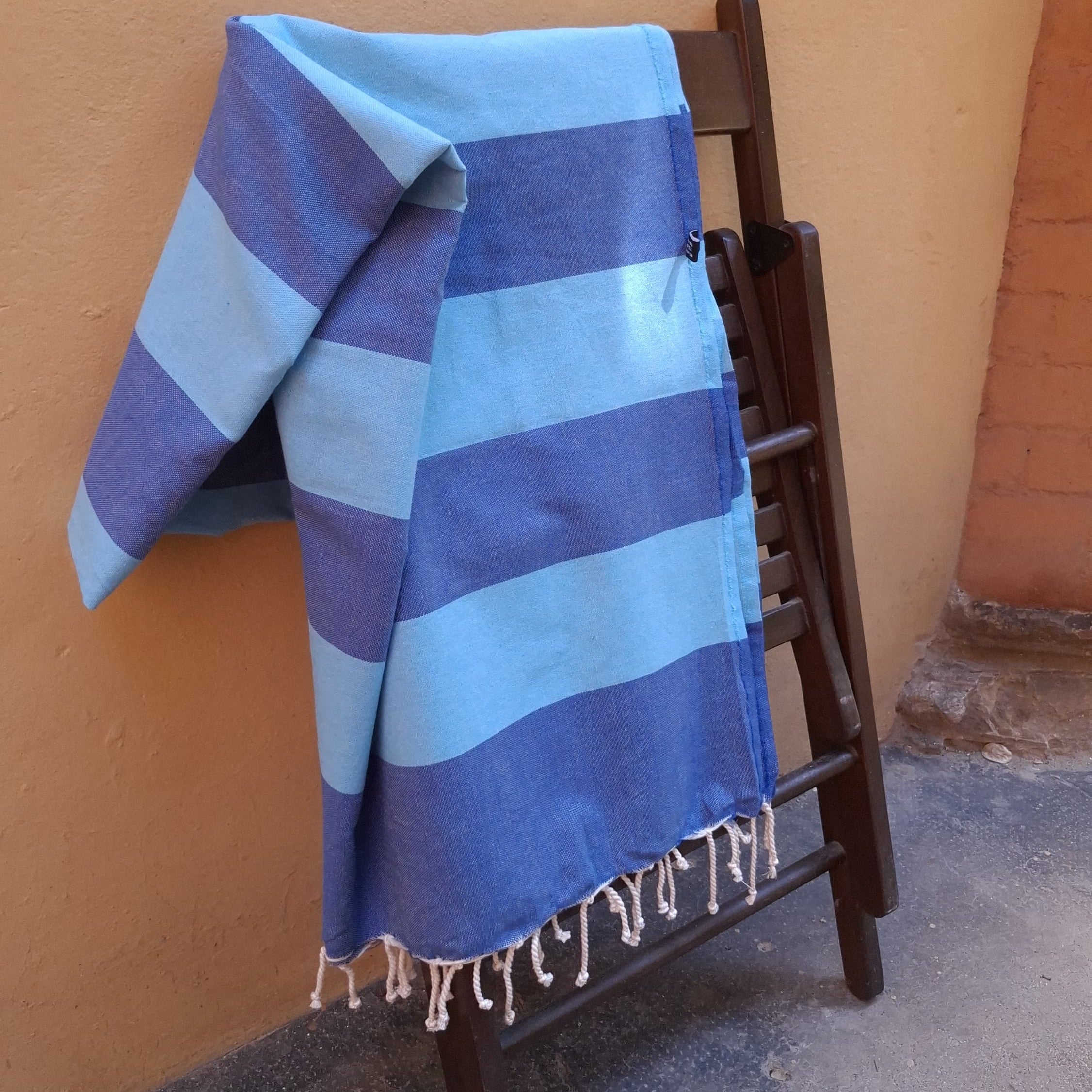 Telo mare Fouta in cotone blu e azzurro steso su una sedia pieghevole in legno all'aperto, contro una parete color ocra.