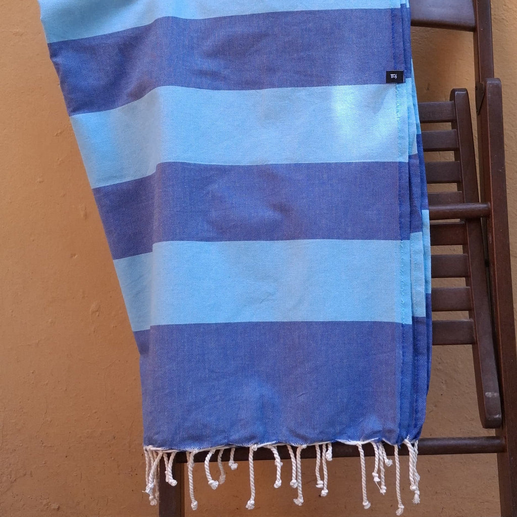 Fouta telo mare a righe blu e azzurre drappeggiato su una sedia in legno con ombre e luce solare naturale."