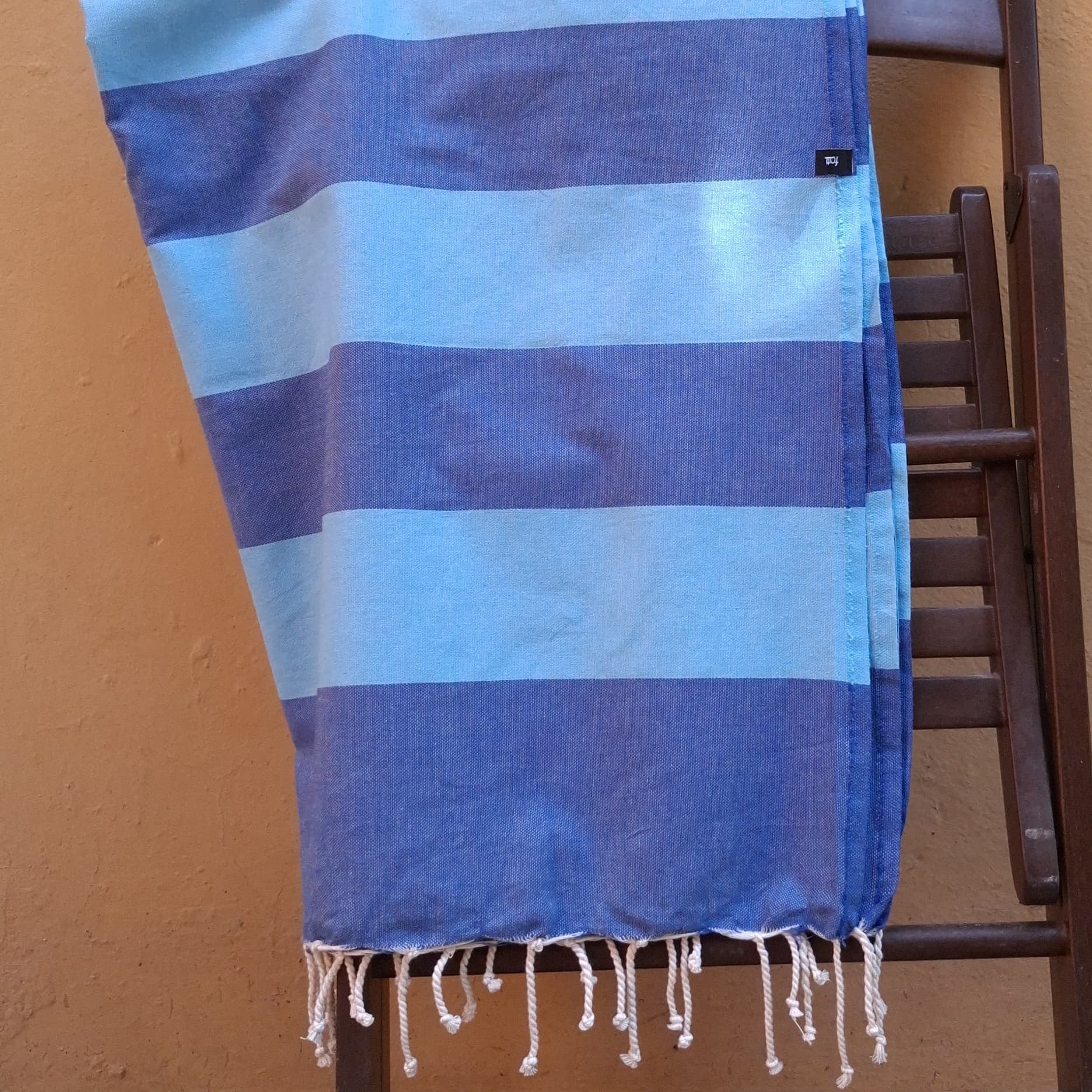 Fouta telo mare a righe blu e azzurre drappeggiato su una sedia in legno con ombre e luce solare naturale."